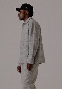 Moški v sivi majici z dolgimi rokavi, ustreznih hlačah in črnim bejzbolaškim klobukom, stoji v profilu pred enobarvno ozadje.