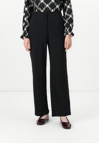 Vrouw in zwarte recht-toe-rechtaan broek, zwart-wit gestreepte blouse en glanzende bordeauxrode platte schoenen tegen een witte achtergrond.