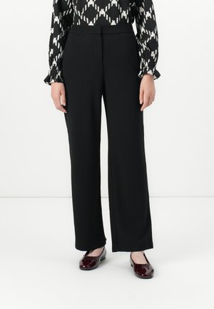 Woman wearing black straight-leg trousers, black and white patterned blouse, and shiny burgundy flat shoes standing against white background.