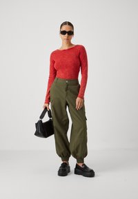 Rode lange mouwen top, olijfgroene cargo broek met zijzakken, zwarte platform schoenen en een kleine zwarte tas. Textuurmateriaal zichtbaar.