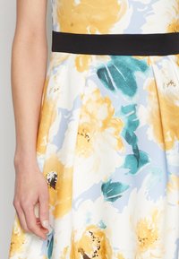 Vestido floral con patrones amarillos, blancos y azules, una cinturilla negra ajustada y una falda acampanada con pliegues. Textura de tela suave.
