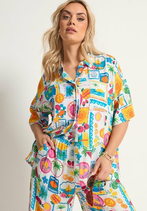 Vrouw met blond haar die een kleurrijk zomerpak met print draagt, bruine zonnebril vasthoudt en met haar hand in haar zak staat tegen een effen achtergrond.