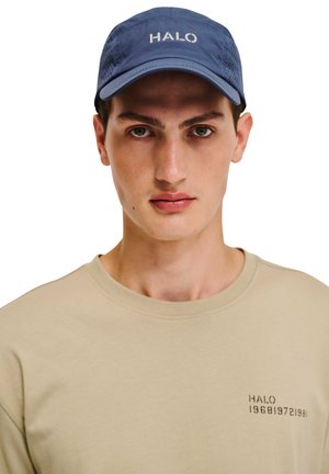 Ung mand iført en navyblå kasket med teksten "HALO" og en beige skjorte med lille sort tekst "HALO 19681972 1981" på brystet.