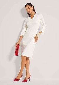 Weißes, figurbetontes Kleid mit V-Ausschnitt und drei Viertelärmeln. Kombiniert mit roten, lackierten Slingback-Heels und einer roten Clutch. Glatte Textur.