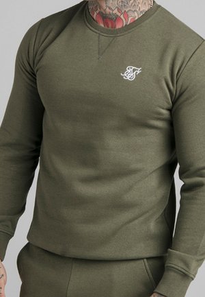Homme portant un sweat-shirt à col rond vert olive avec un logo blanc brodé sur la poitrine gauche, montrant un cou et une main tatoués.