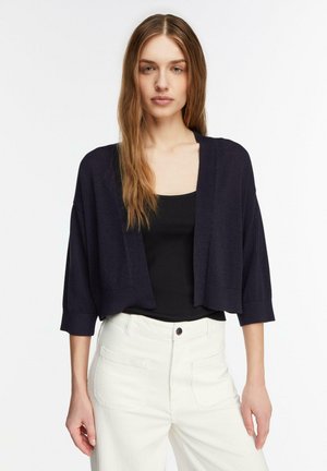 Femme aux longs cheveux portant un cardigan noir court sur un haut noir et un pantalon blanc taille haute, debout devant un fond blanc uni.