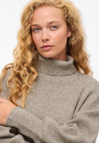 Pilkas ribotas golfas megztinis ilgomis rankovėmis, pasižymintis minkšta tekstūra ir laisvu siluetu. Išskirtinis dėl aukšto kaklo ir šiltos spalvos.