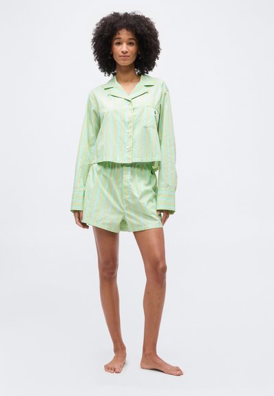 Γυναίκα με σγουρά μαλλιά, ντυμένη με ανοιχτό πράσινο πουκάμισο με ρίγες και μακριά μανίκια, και ταιριαστό σορτς, στέκεται ξυπόλυτη μπροστά σε απλό φόντο.