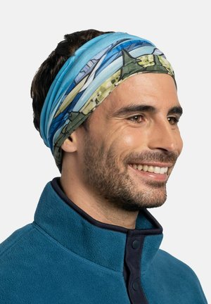 Hombre sonriendo con una chaqueta de forro polar azul y una diadema multicolor azul y verde con patrón sobre un fondo blanco.
