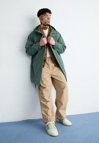 Giacca con cappuccio verde, maglietta beige, pantaloni beige. Sneakers bianche con accenti verdi. Tessuto morbido, design casual, vestibilità rilassata.