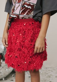 Červená sukňa s textúrou pripomínajúcou perie a flitrami; priliehavý pás s elastickou páskou. Kombinovaná s oversize tričkom s grafikou a zdobeným flitrami.