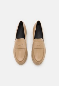 Mocassins en cuir beige avec un design fermé, bout arrondi et petit détail d'accent à l'avant. Semelle intérieure noire texturée visible.