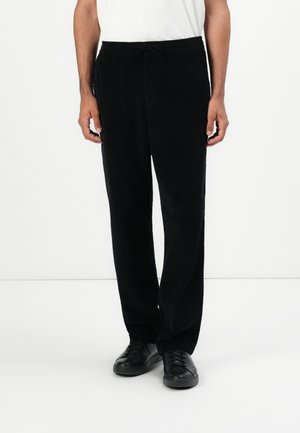 Man in zwarte corduroy broek met een trekkoord in de taille en zwarte sneakers, staand tegen een effen witte achtergrond.