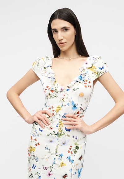 Vrouw met lang donker haar, gekleed in een witte bloemenjurk met geplooide mouwen, poseert met handen in de zij tegen een effen achtergrond.