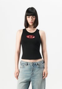 Schwarzes, kurzes Tanktop mit einem runden roten und weißen Logo in der Mitte; kombiniert mit hellblauen, weiten Jeans. Glattes Material, figurbetonter Schnitt.