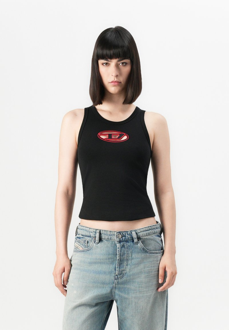 Schwarzes, kurzes Tanktop mit einem runden roten und weißen Logo in der Mitte; kombiniert mit hellblauen, weiten Jeans. Glattes Material, figurbetonter Schnitt.