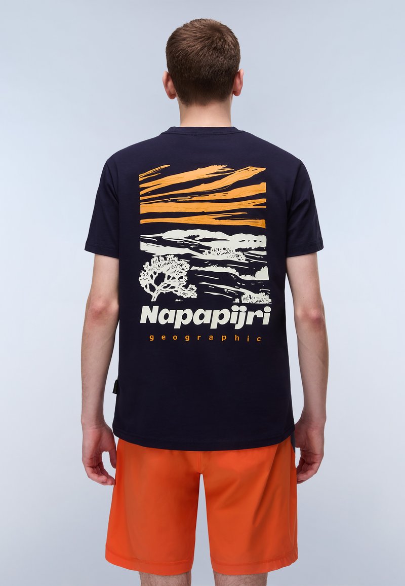 Camiseta azul marino con un gráfico de paisaje colorido y el texto "Napapijri geographic" en la parte de atrás. Combinada con pantalones cortos naranjas.