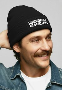 Black ribbed beanie with a rectangular white patch featuring the text "HÄNDHERUM GLÜCKLICH." Soft texture, fitted design.