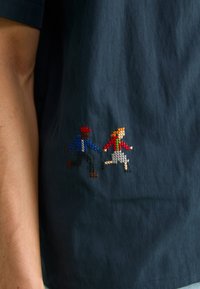 To personer brodert med korssting, en person med rød cap og blå jakke som holder hender med en person i rød genser og grå skjørt, på mørkt stoff.