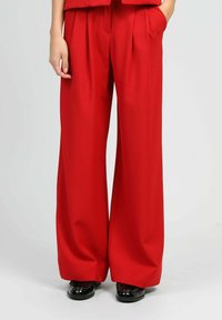 Pantalons larges rouges en tissu lisse, dotés de plis et de poches latérales. Associés à des chaussures noires brillantes.