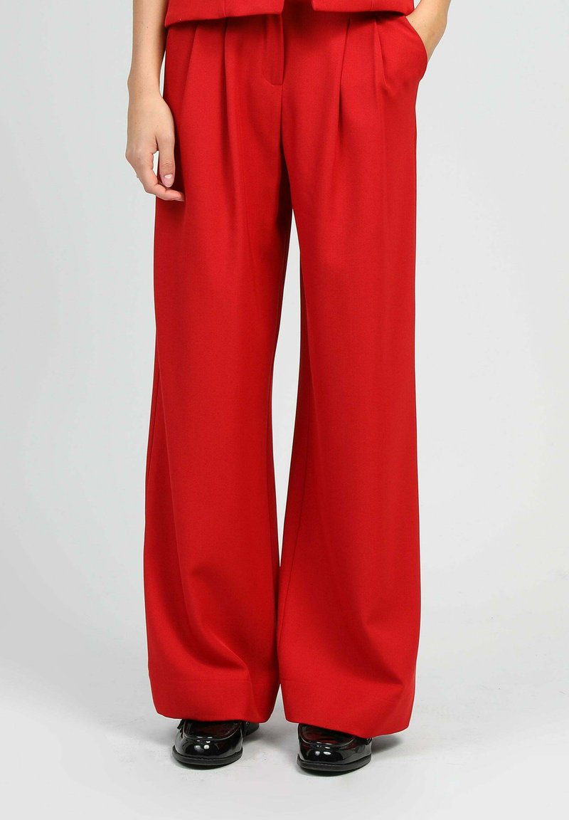 Pantalons larges rouges en tissu lisse, dotés de plis et de poches latérales. Associés à des chaussures noires brillantes.