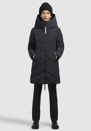 Manteau matelassé marine à col haut, design en chevrons et fermeture éclair à l'avant. Associé à un pantalon noir et un bonnet en tricot.