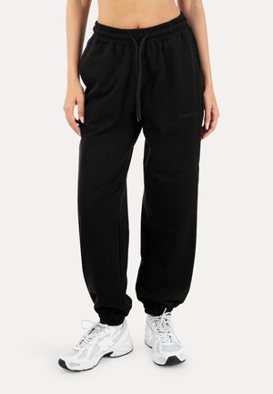 Osoba ubrana w czarne dresowe spodnie z elastycznym pasem i sznurkiem oraz białe sportowe buty, stojąca na szarym tle.