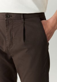 Brown cotton trousers featuring a front pleat, side pockets, and a single button closure at the waist. Smooth texture and tailored fit.