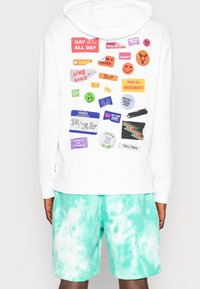 Vit hoodie med färgglada lappar som främjar LGBTQ+-rättigheter, pronomen, Black Lives Matter och social rättvisa, bärs med gröna tie-dye-shorts.