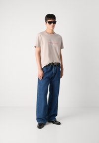 T-shirt en coton beige avec le logo "Calvin Klein Jeans" en rouge, associé à un jean en denim bleu ample et des chaussures en cuir noires. Lunettes de soleil noires portées.