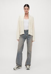 Χοντρή κρεμ καμπαρντίνα με πλέξη καλωδίου και ψηλό γιακά, ανοιχτό μπροστά, με δύο μπροστινές τσέπες, σε συνδυασμό με ανοιχτό μπλε ψηλόμεσα τζιν και αθλητικά παπούτσια.