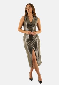 Abito avvolgente argento metallico con uno spacco fino al ginocchio, design senza maniche e texture liscia. Abbinato a scarpe nere con tacco alto.