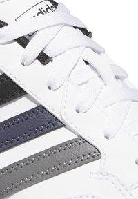 Sneaker bianca con lacci bianchi, righe zigzag cucite in nero, blu navy e grigio, e un logo Adidas parzialmente visibile sulla linguetta.