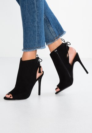 Czarne zamszowe botki na wysokim obcasie z odkrytymi palcami, wycięciami po bokach, suwakiem z tyłu i ozdobą w formie kokardy. Charakteryzują się eleganckim krojem i zaostrzonym noskiem.