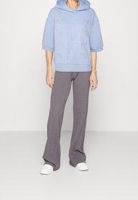 Osoba oblečená v světle modré mikině s krátkým rukávem a šedých širokých kalhotách s jemným vzorem, v bílé sneakers, stojící na pozadí bez detailů.