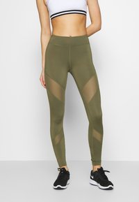 Leggings verde oliva con paneles de malla en un patrón de chevron. Diseño de cintura alta, textura suave, combinados con zapatillas deportivas negras.