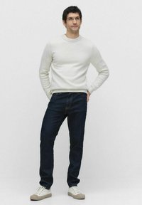 Ljusgrå stickad tröja med rund halsringning, tillsammans med mörkblå jeans och vita sneakers, stående mot en enfärgad vit bakgrund.