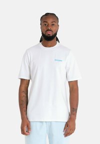 T-shirt blanc en coton à manches courtes, avec un petit logo bleu sur la poitrine gauche. Texture lisse et coupe décontractée.