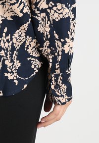 Košile s květinovým vzorem s tmavě námořnickým pozadím a světlými růžovými květy. Má manžety na knoflíky a hladkou texturu látky.