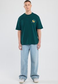 T-paita tummanvihreää puuvillaa, jossa lyhyet hihat, pyöreä pääntie ja etupuolella keltainen ja valkoinen logo. Yhdistetty vaaleansinihin leveisiin farkkuihin.