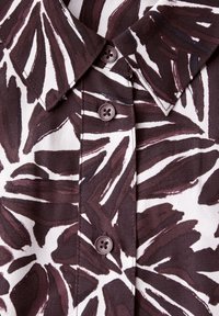 Chemise à manches courtes avec un fond blanc et un motif de feuilles bordeaux. Elle possède un col pointu et quatre boutons bordeaux.