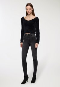 Pullover nero peloso corto con scollatura ampia, abbinato a jeans attillati grigio scuro. La modella indossa stivali neri con tacco alla caviglia. Sfondo semplice.