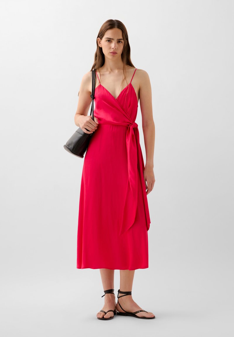 Vestito lungo rosso con design a pettine, spalline sottili e un dettaglio a nodo in vita. Indossato con sandali neri e una piccola borsa nera.