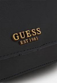 Černá kožená peněženka s zlatým logem "GUESS" a rytím "EST 1981". Hladký povrch s neat švy a minimalistickým designem.