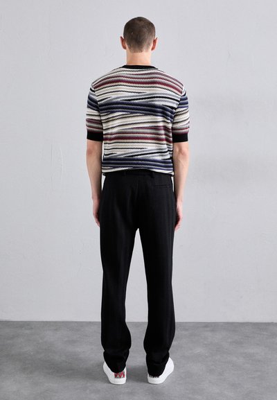 Pīnēta svītraina džemperis bēšā, sarkanā un zilā krāsā ar īsām piedurknēm, saskaņots ar melniem teksturētiem biksēm un baltām sporta apaviem ar rakstainiem akcentiem.