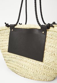 Πλεκτή τσάντα από ψάθα με μαύρη δερμάτινη λεπτομέρεια. Διαθέτει πλεξούδες μαύρες χελιδονοούρες και μια διακοσμητική μαύρη ετικέτα με λεπτομέρεια λογότυπου σε περφορέ.