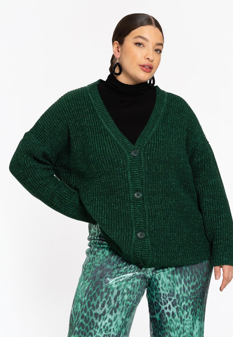 Dunkelgrüner, gestrickter Cardigan mit V-Ausschnitt, oversized Passform, fünf Knöpfen und gerippter Textur, kombiniert mit glänzenden grünen Leopardenmusterhosen.