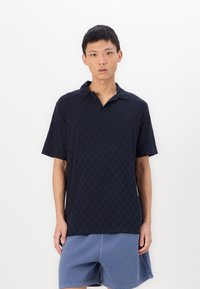 Ναυτικό καρό πόλο πουκάμισο κοντομάνικο με γιακά, συνδυασμένο με ανοιχτό μπλε σορτς. Το ύφασμα φαίνεται υφασμένο, με καθημερινή εφαρμογή.