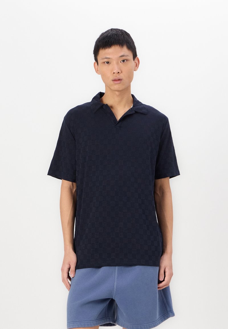 Ναυτικό καρό πόλο πουκάμισο κοντομάνικο με γιακά, συνδυασμένο με ανοιχτό μπλε σορτς. Το ύφασμα φαίνεται υφασμένο, με καθημερινή εφαρμογή.