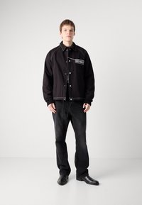 Giacca nera in cotone con stitching bianco a contrasto e logo "HUGO". Indossata sopra una camicia nera e abbinata a jeans scuri e scarpe nere.